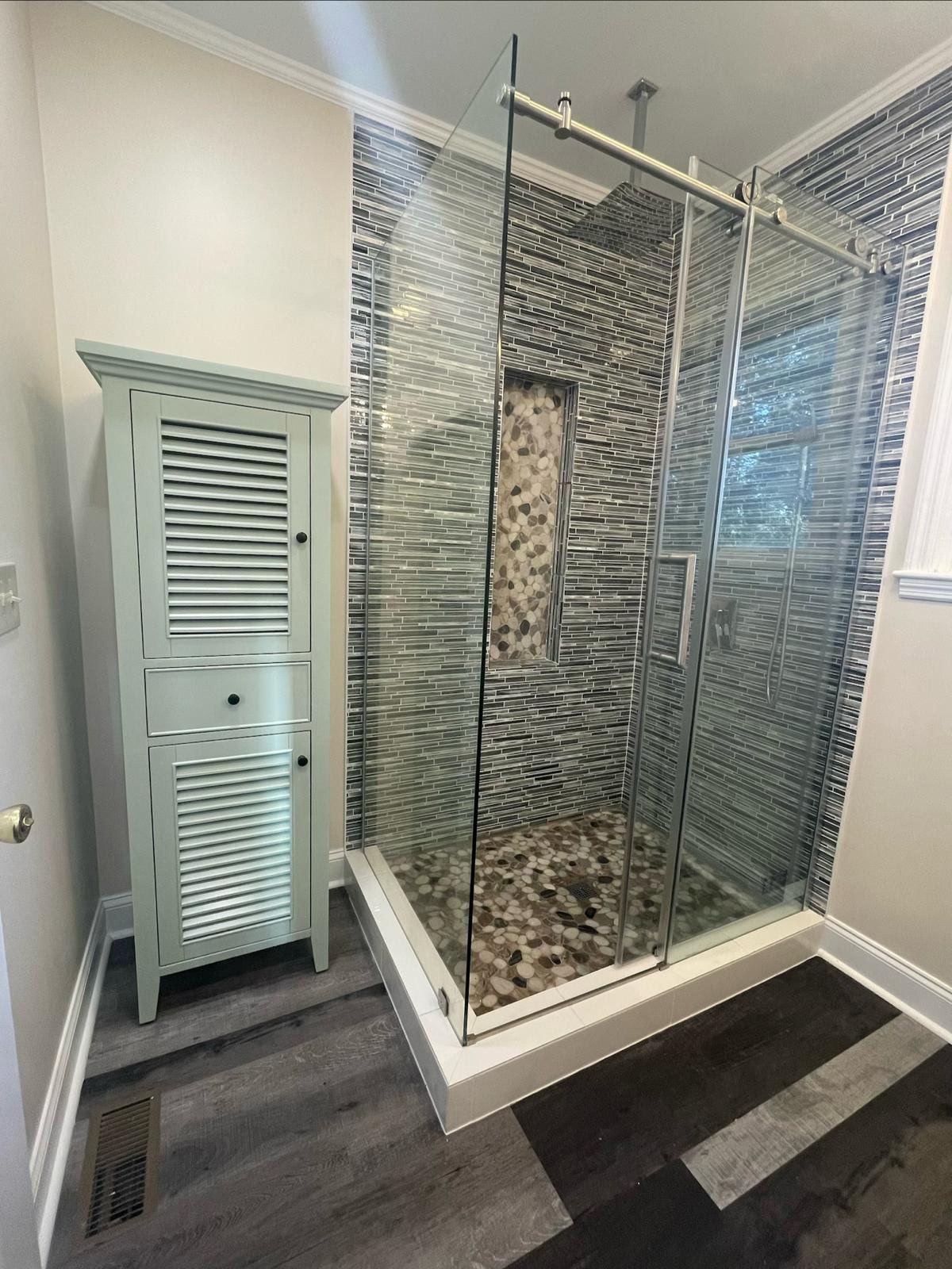 Glass-enclosed corner shower with stone tile wall, next to a pale green louvered cabinet in a bathroom