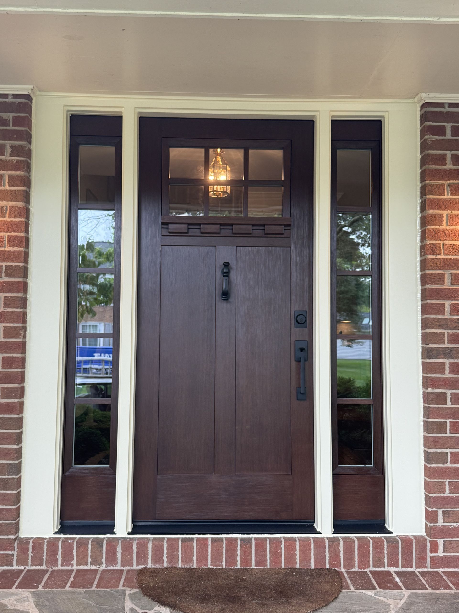 Brown wooden front door with sidelights, brick facade, and welcome mat.