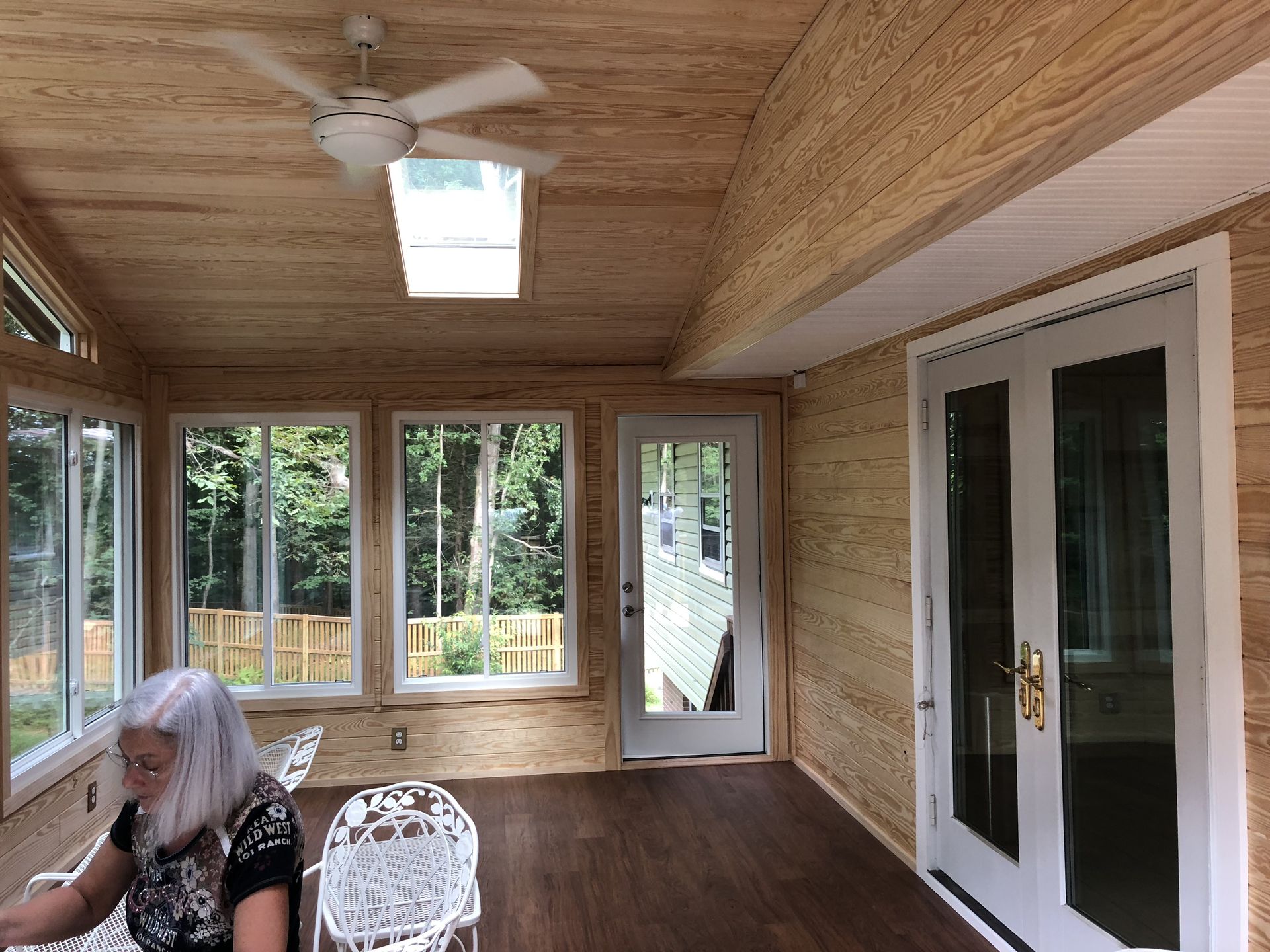 Sunroom with wood paneling, windows, a ceiling fan, and a woman seated.