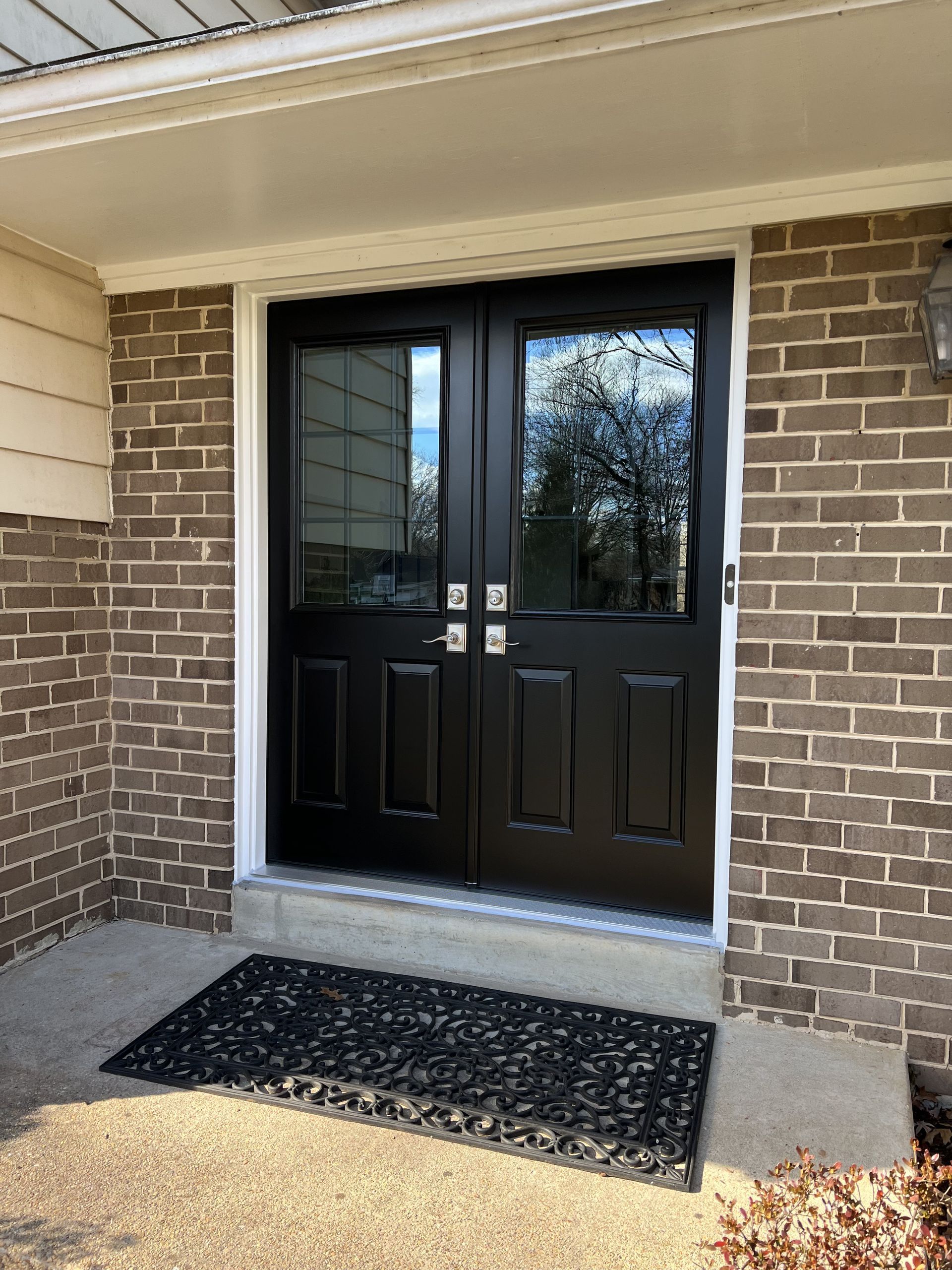 Black double doors with glass panels and white trim, set in a brick entryway with a welcome mat.