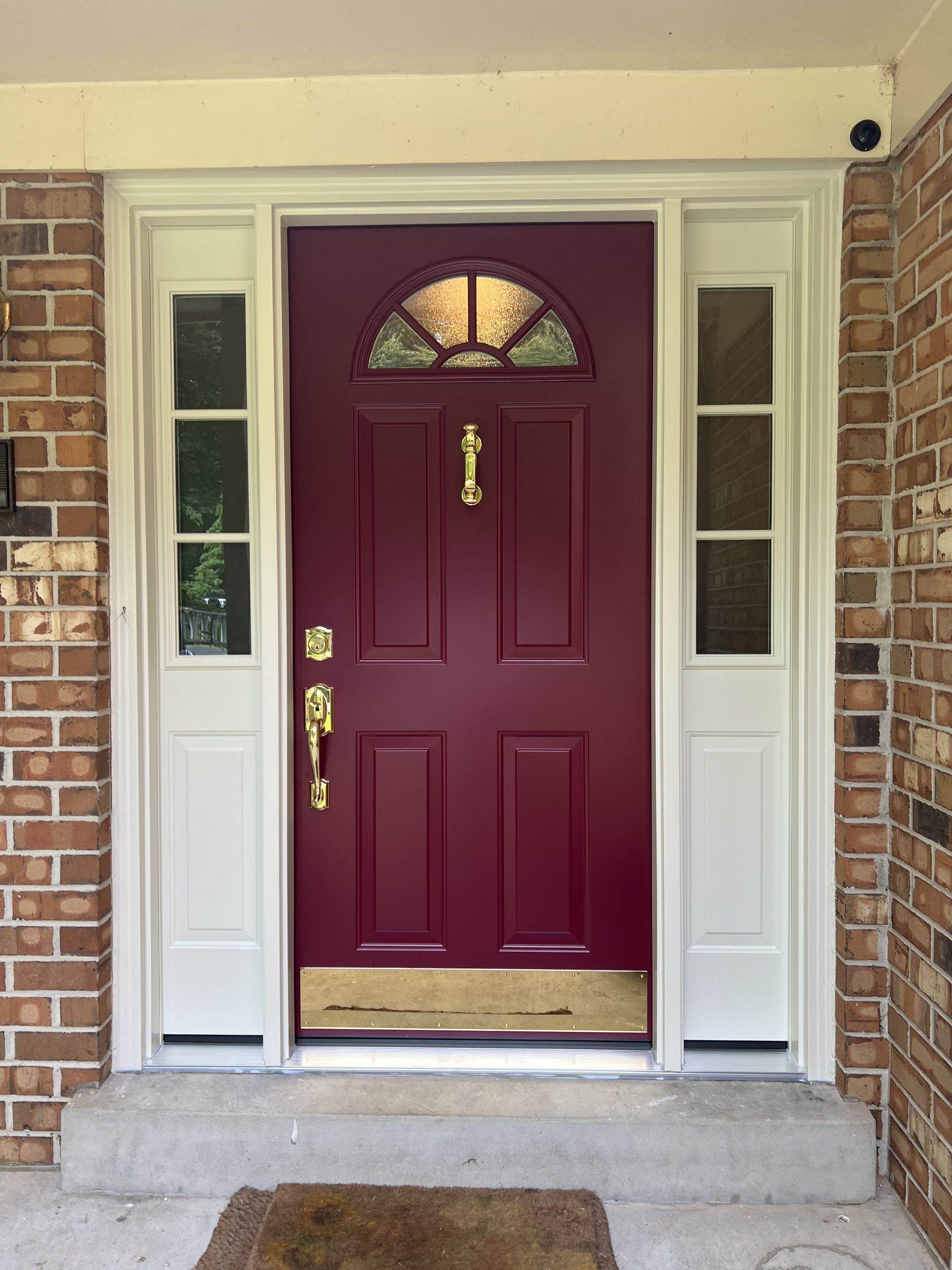 Maroon front door with sidelights, brass accents, and a decorative top window. Set in a brick house.