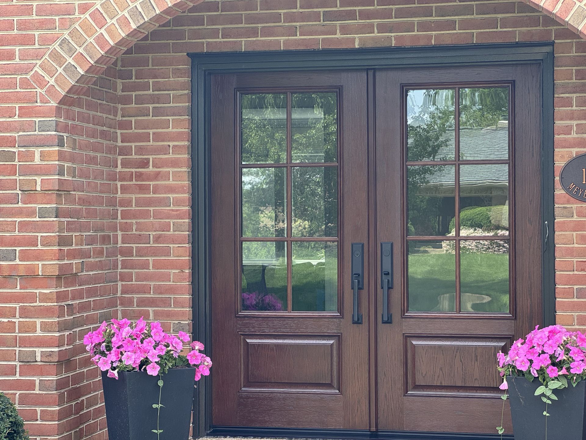 Double wooden doors with glass panels and black handles, flanked by pink flower planters, set against a red brick facade.