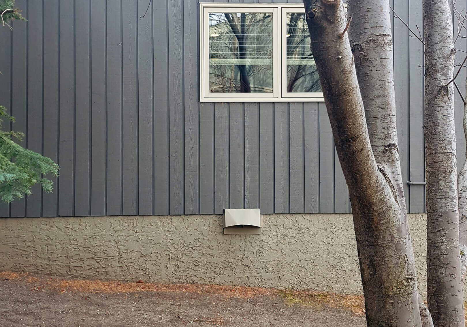 A gray house with a window and trees in front of it.