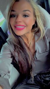 A person with long blonde hair and bright pink lipstick smiling while sitting in a car.