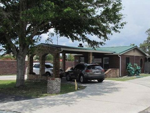 A single-story brick building with a covered arched carport, featuring a gray SUV parked under it on a sunny day.