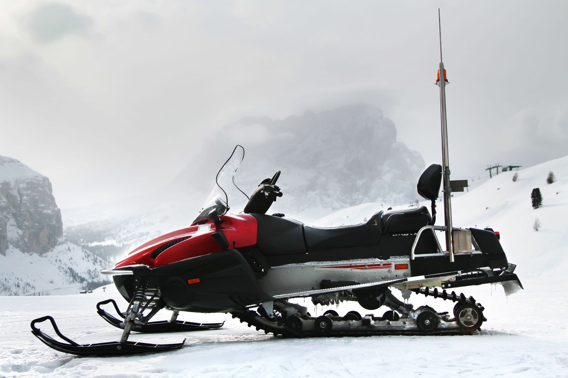 Red and black snowmobile with antenna in snowy mountain landscape.