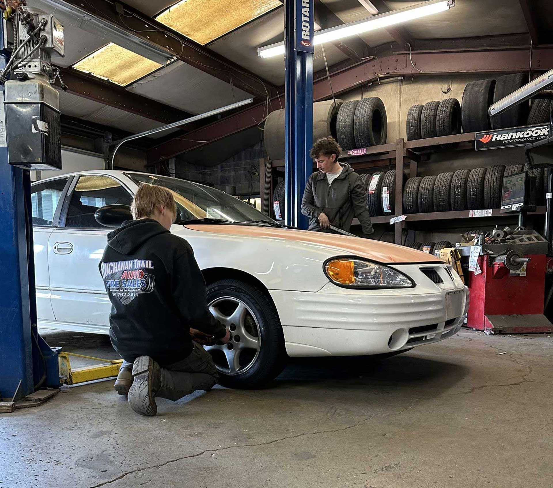 Two mechanics work on a white car in a garage with tires on a shelf. One kneels by the wheel.
