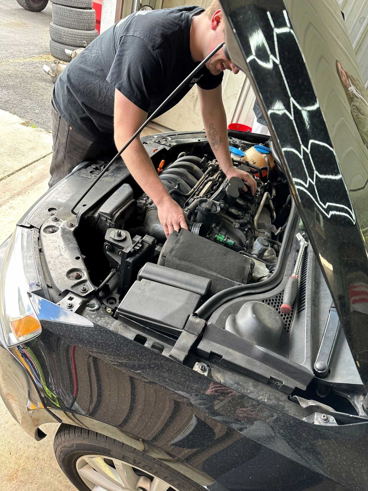 Mechanic working on a black car engine with hood open, outdoors.