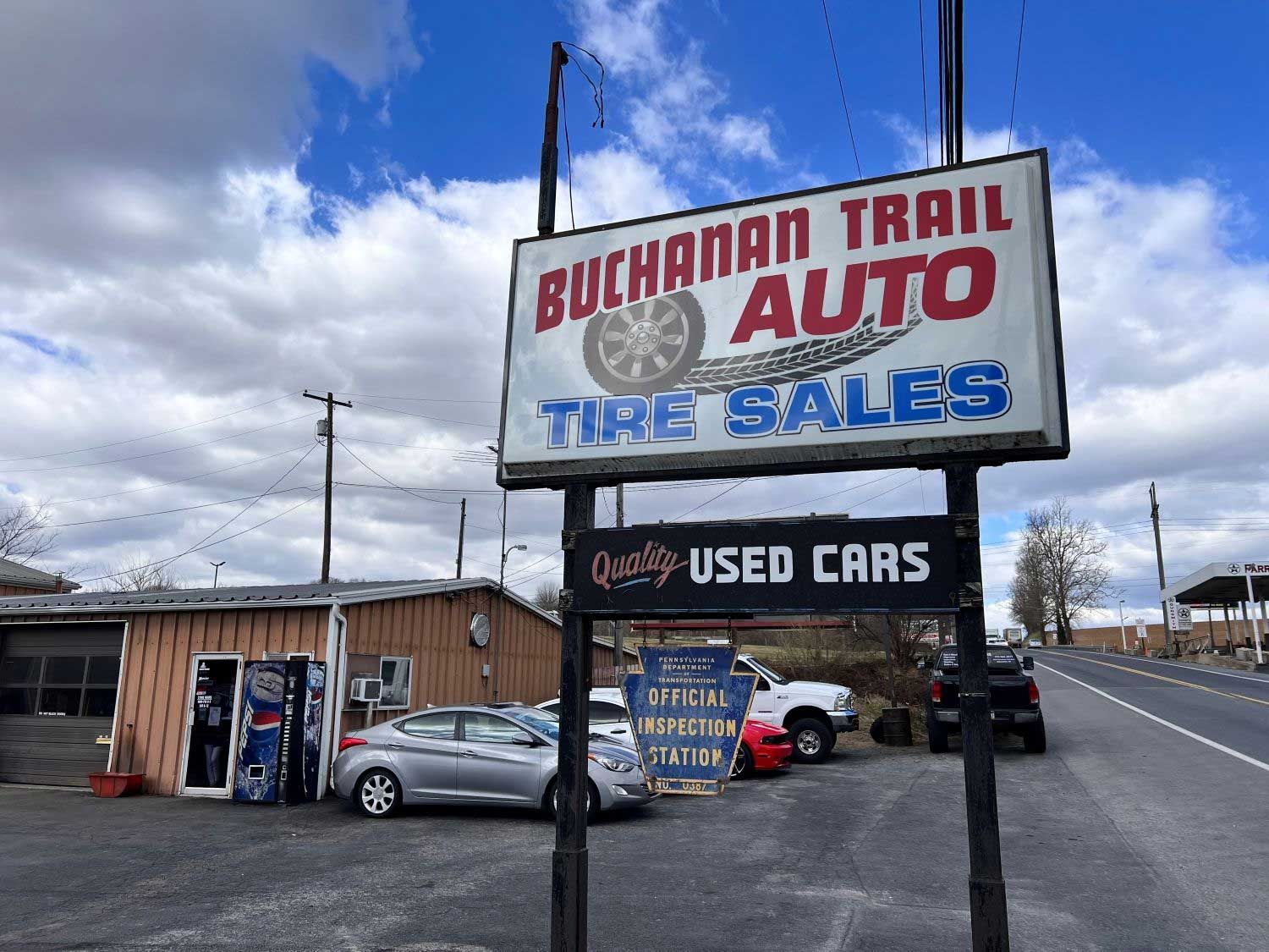 Buchanan Trail Auto sign with tire sales, used cars. Cars parked in front of small garage under partly cloudy sky.