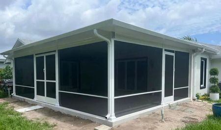 Screened-in porch addition with white framing, dark screening, and a white roof against a cloudy sky.