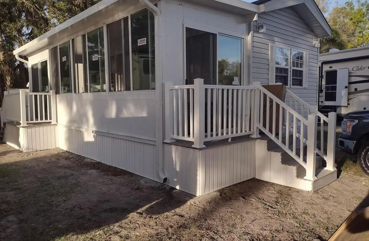 White-sided house with deck and porch. Steps lead up to the door. Sunlight, gravel yard, and RV nearby.