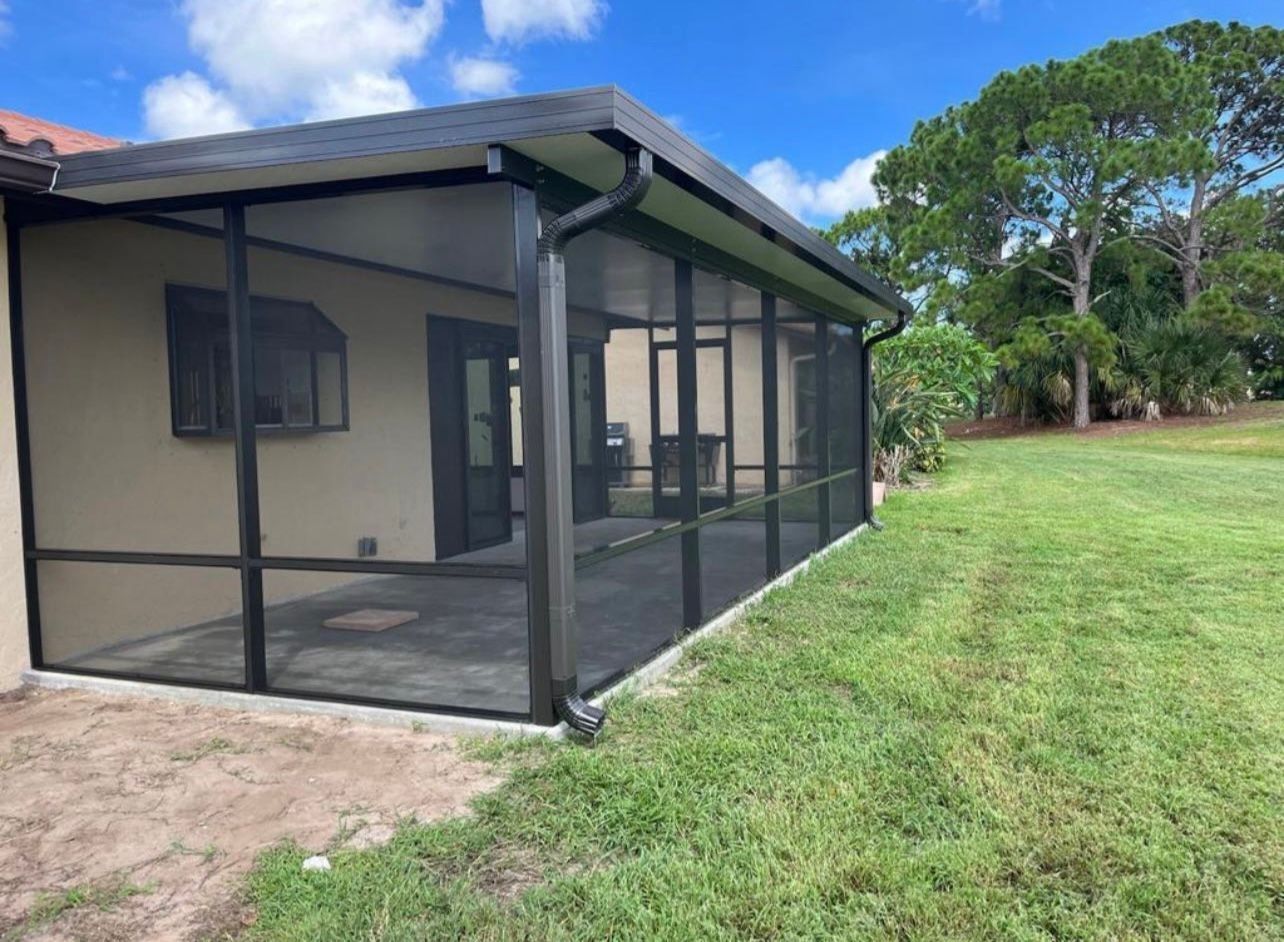 Screened-in patio with black framing and dark screens, attached to a beige house, overlooking a grassy yard under a blue sky.