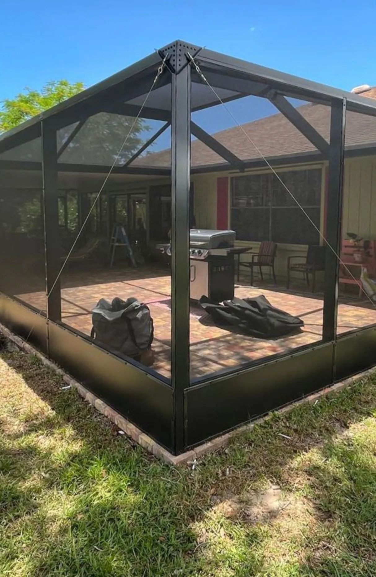 Black screened-in patio enclosure with a grill and patio furniture on a brick surface. Green lawn surrounds it.