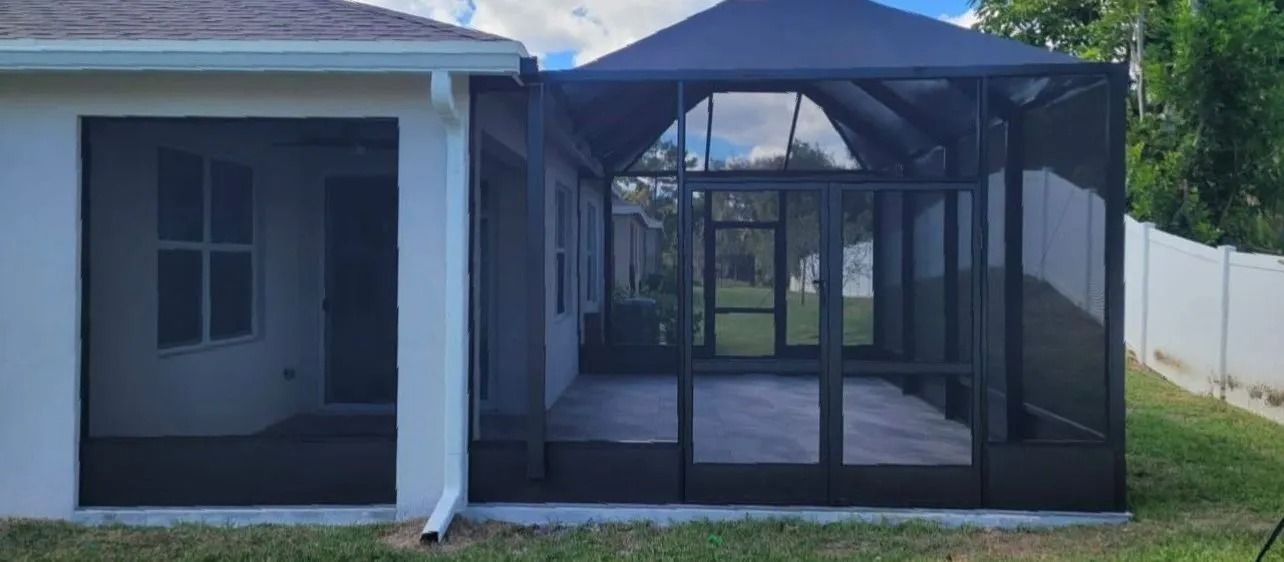A screened patio attached to a house with a concrete floor, surrounded by a yard and white fence.