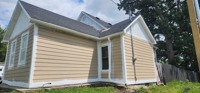 Tan house with white trim, dark roof, and a grassy yard under a cloudy sky.