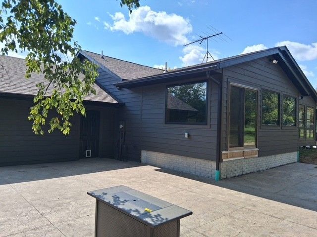 Dark-sided house with large windows, an open patio, and a fire table, under a blue sky.