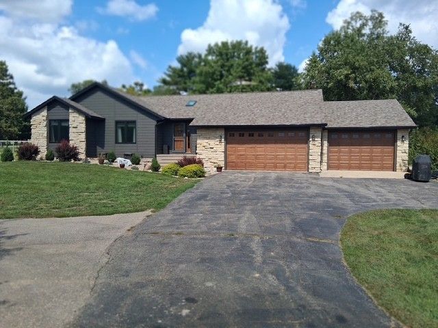 Ranch-style house with stone and gray siding, brown garage doors, and a paved driveway under a blue sky.