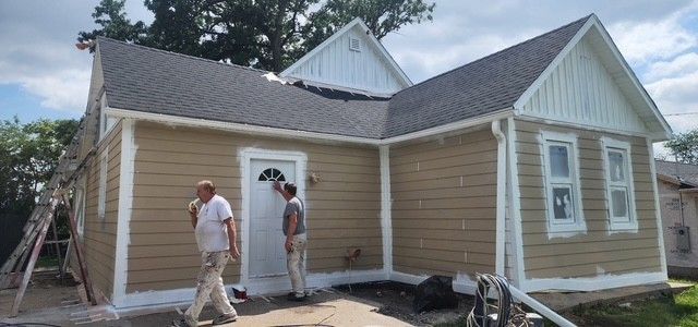 House exterior being painted by two people. Building has beige siding, white trim, and a dark roof.