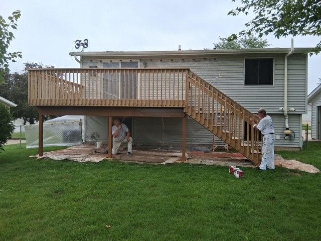 Two people painting under a wooden deck attached to a two-story house. They wear white suits. Green grass surrounds.
