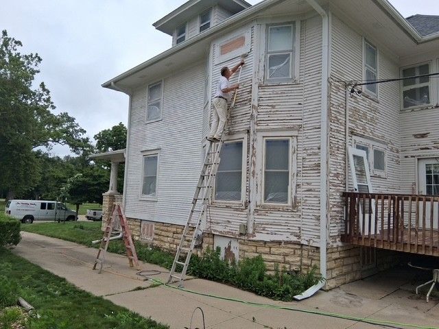 Person on a ladder scraping paint off a two-story white house with peeling siding.