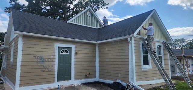 Two people painting a tan house with white trim. One on the roof, the other on a ladder.