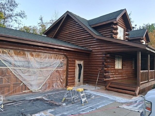 Brown log cabin being painted with garage door covered in plastic and tarps on ground.