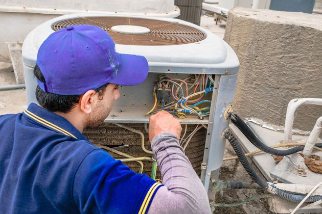 Technician wiring an outdoor air conditioner unit beside a building wall