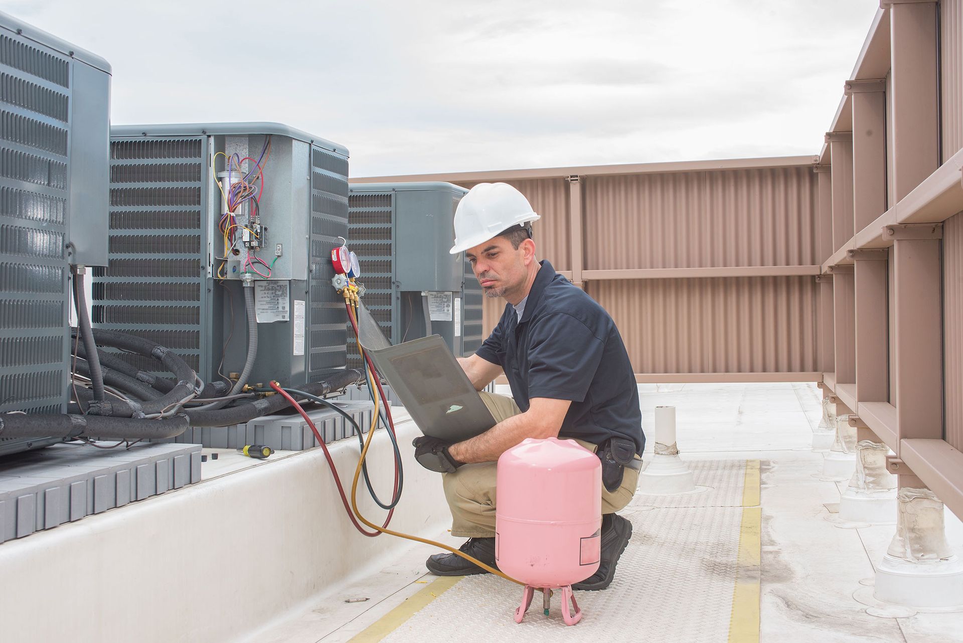 Technician servicing rooftop HVAC units beside a pink gas cylinder and laptop