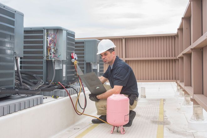 Technician servicing rooftop HVAC units beside a pink gas cylinder and laptop