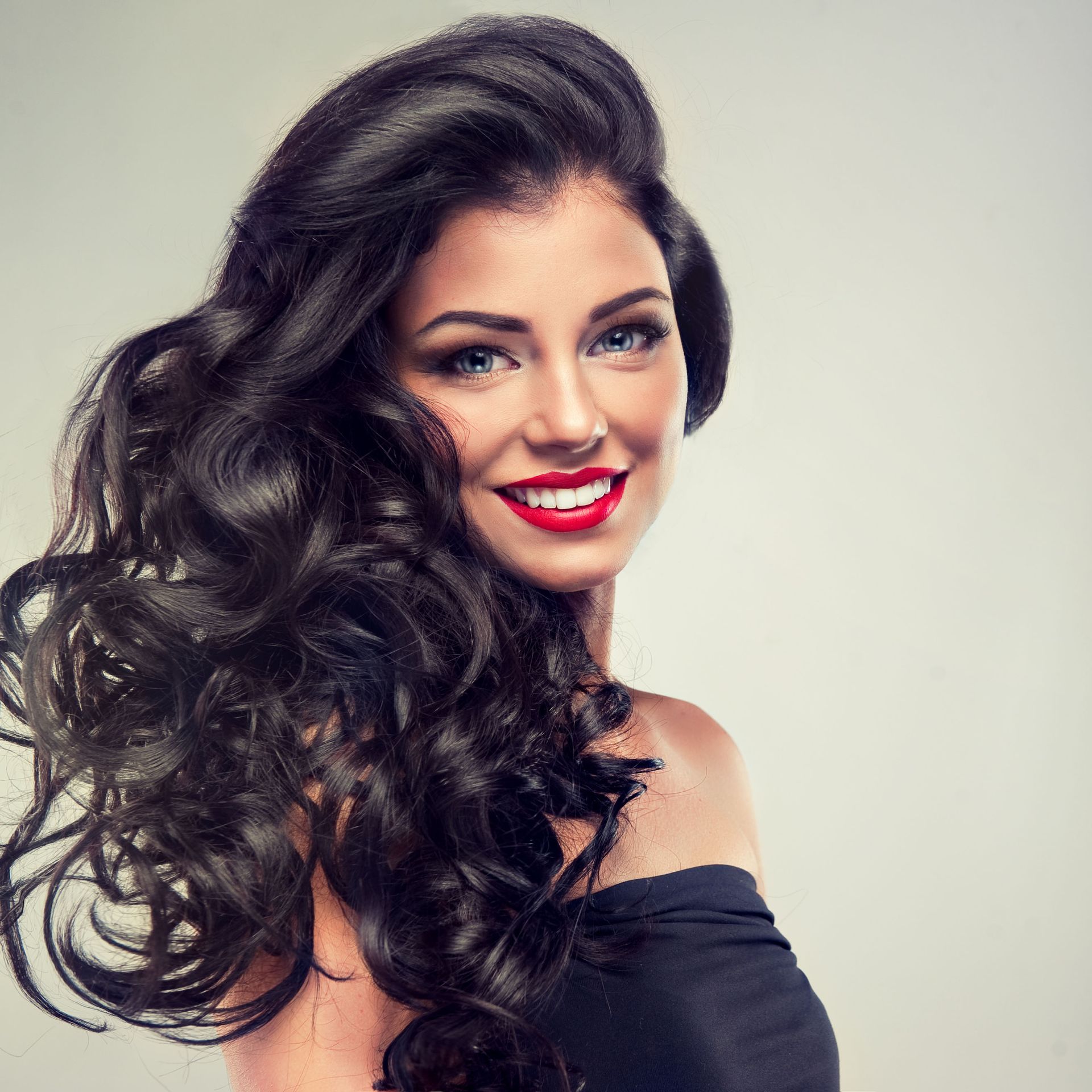 Woman with dark, wavy hair, smiling and looking at the viewer; wearing red lipstick and a black top.