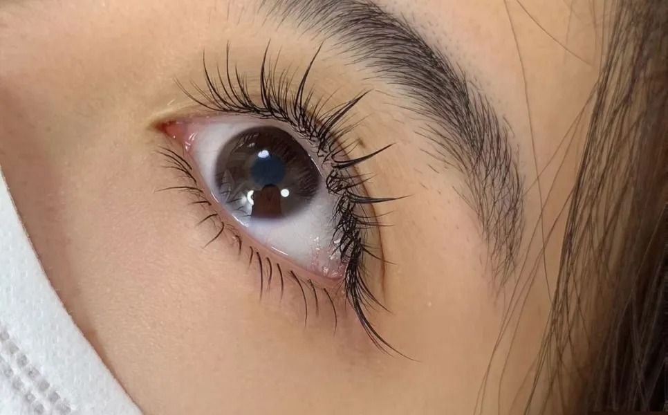 Close-up of an eye with long, curled eyelashes and a defined eyebrow; visible white face mask.