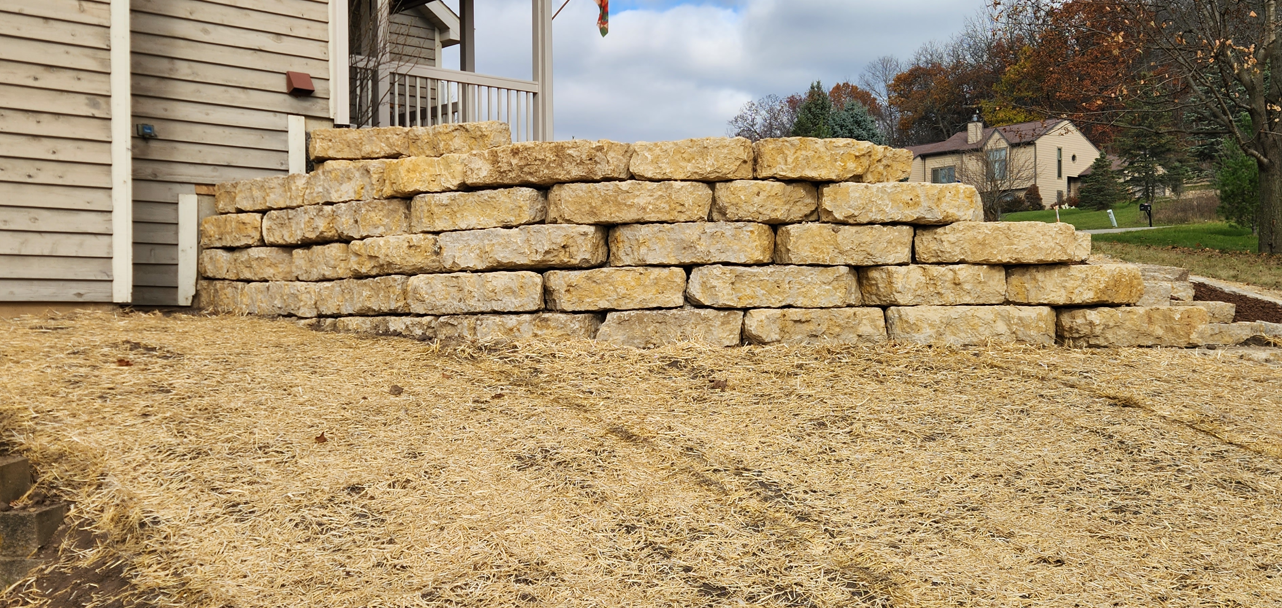 Stacked tan retaining wall blocks beside a house on a gravel slope under a cloudy sky