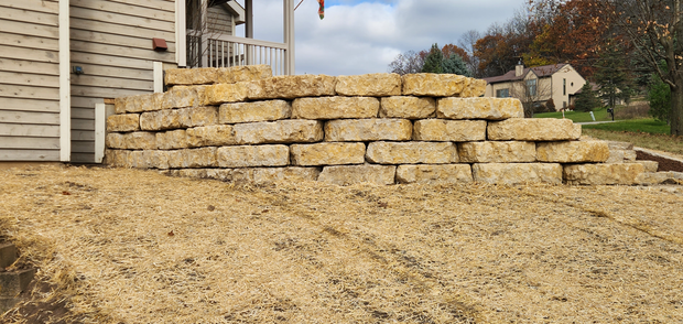 Stacked tan stone retaining wall beside a beige house on a gravel slope