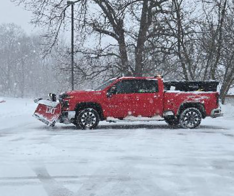 Red pickup truck with a snowplow clearing a snowy road in a winter storm