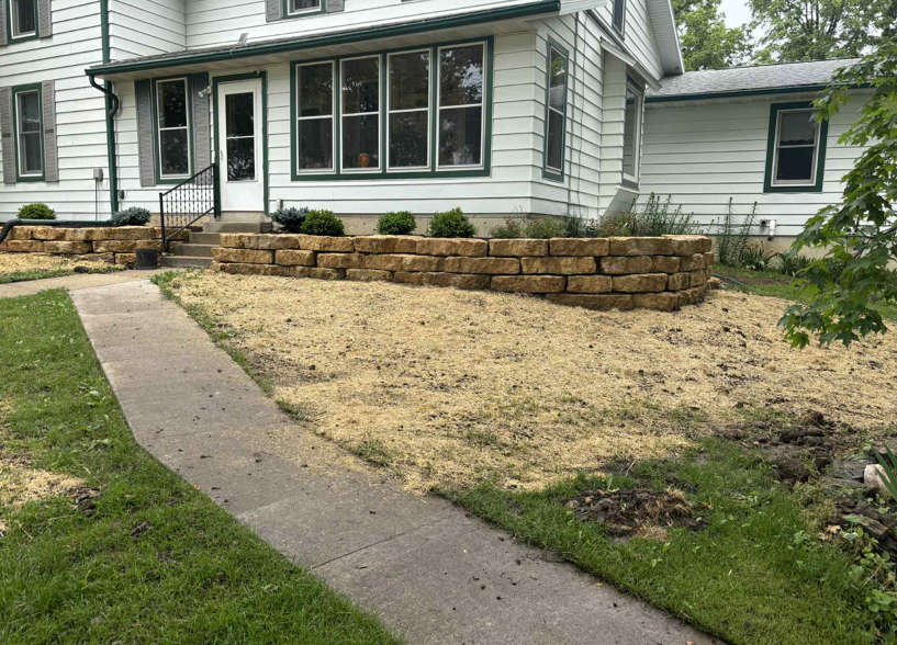 Front yard with a curved walkway, dry lawn, and stacked stone retaining wall beside a white house.