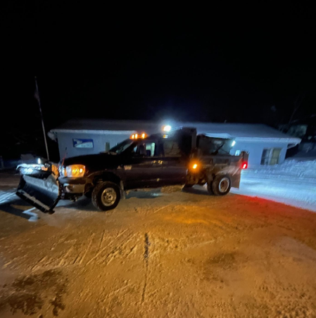 Tow truck with plow attached parked on a snowy road at night in front of a building