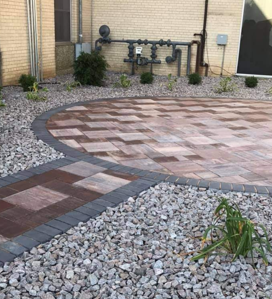Paved patio with brick border, gravel landscaping, and utility pipes against a beige wall.