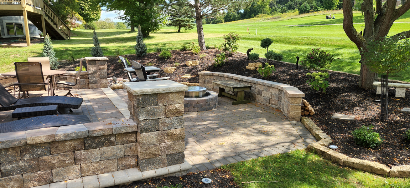 Stone patio with built-in fire pit and seating in a landscaped backyard garden, surrounded by trees and lawn