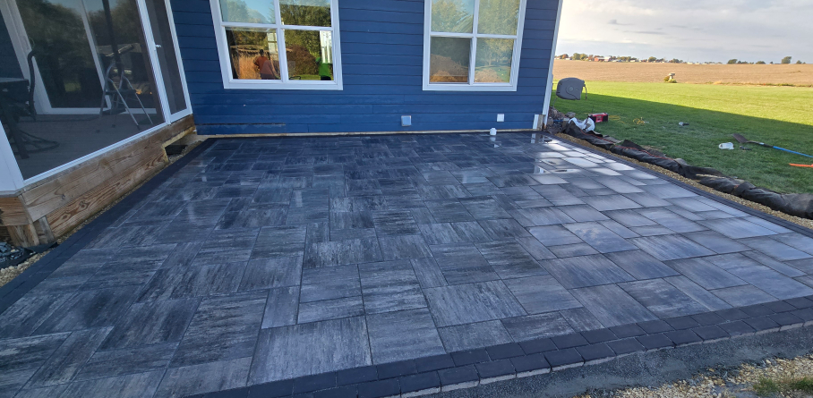 Dark gray stone patio beside a blue house with windows and a glass door, bordered by lawn and landscaping
