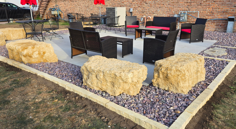 Outdoor patio seating area with black chairs, tables, red umbrellas, and large stone benches on gravel.