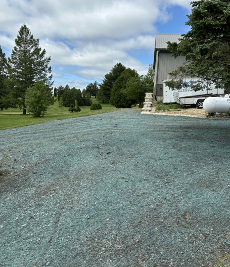 Gravel driveway beside a house, with trees, lawn, and a white boat or trailer under a cloudy sky.
