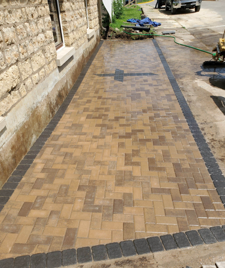 Long paved walkway beside a stone wall, with dark border bricks and construction materials at the far end.
