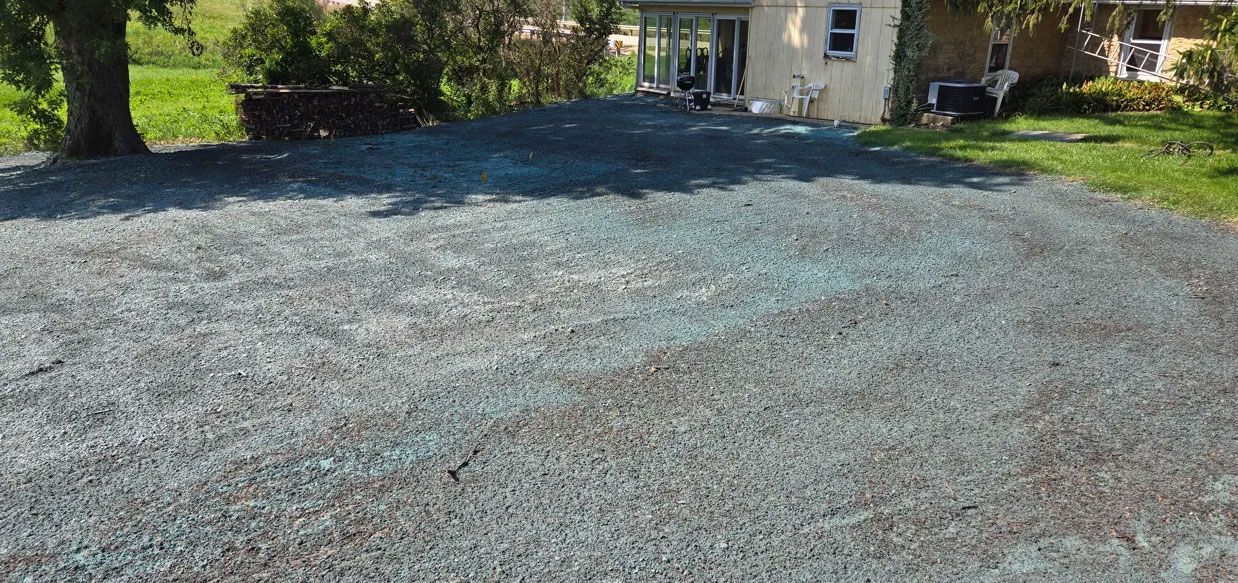 Gravel driveway leading to a house with a black carport and green lawn on a sunny day