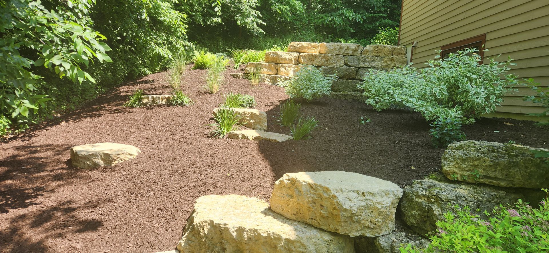 Mulched garden bed with shrubs, large rocks, and a stone pathway beside a house