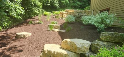 Mulched garden path with large stone steps beside a house, bordered by green shrubs and trees