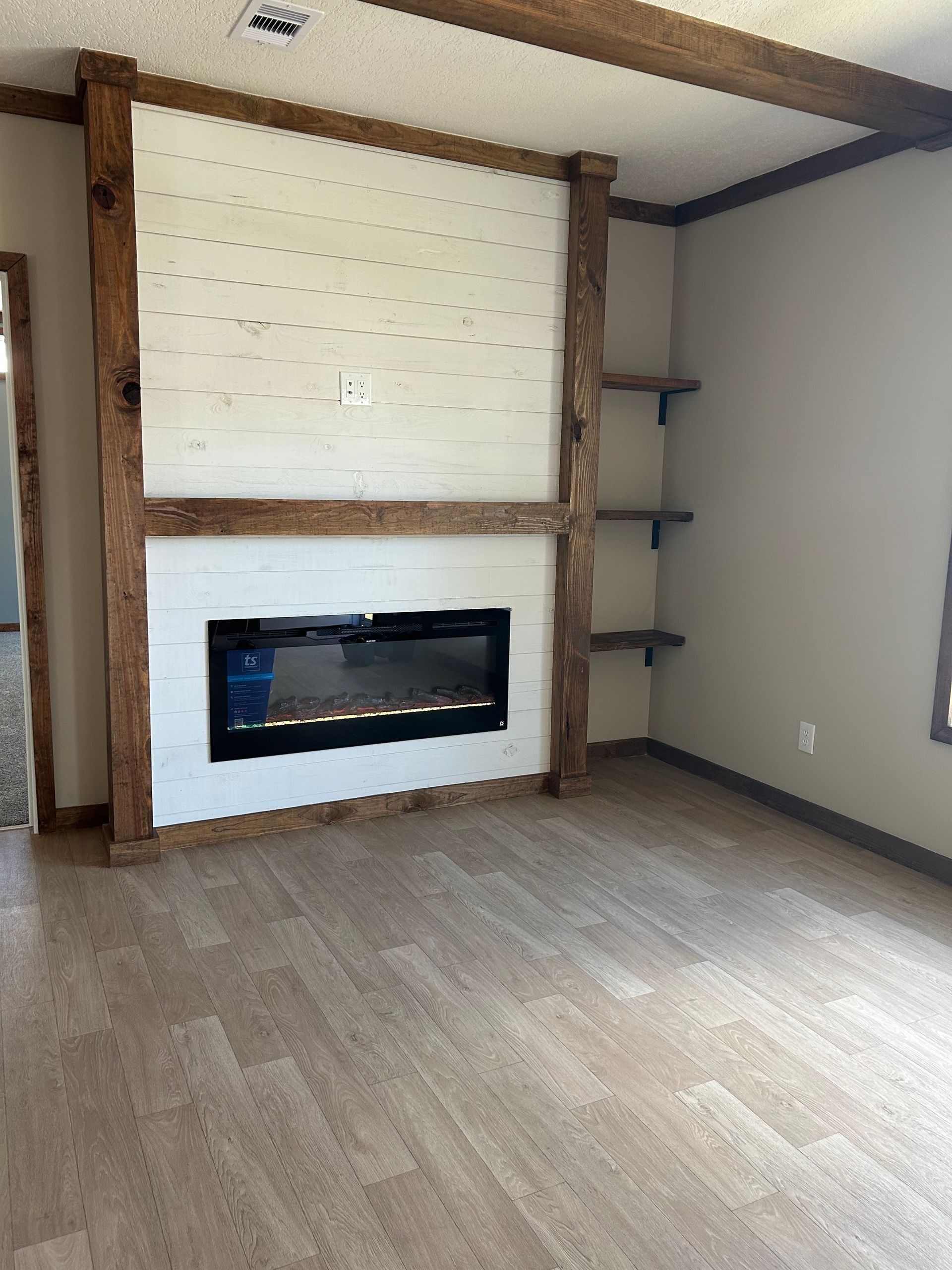 Living room with fireplace, white brick wall, wooden beams, shelves, and beige flooring.