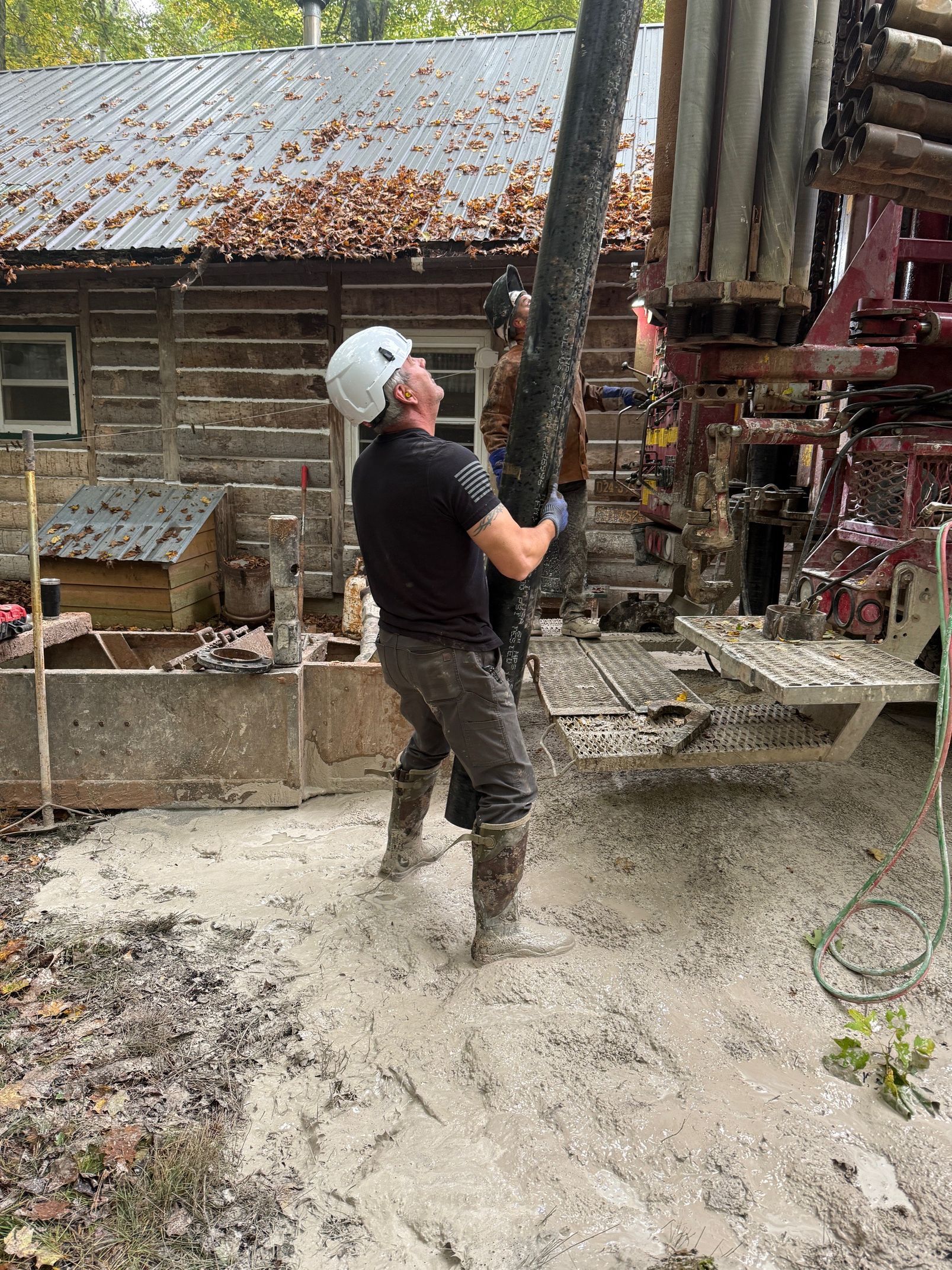 Worker in a helmet shoveling wet concrete beside a drilling rig at a muddy construction site