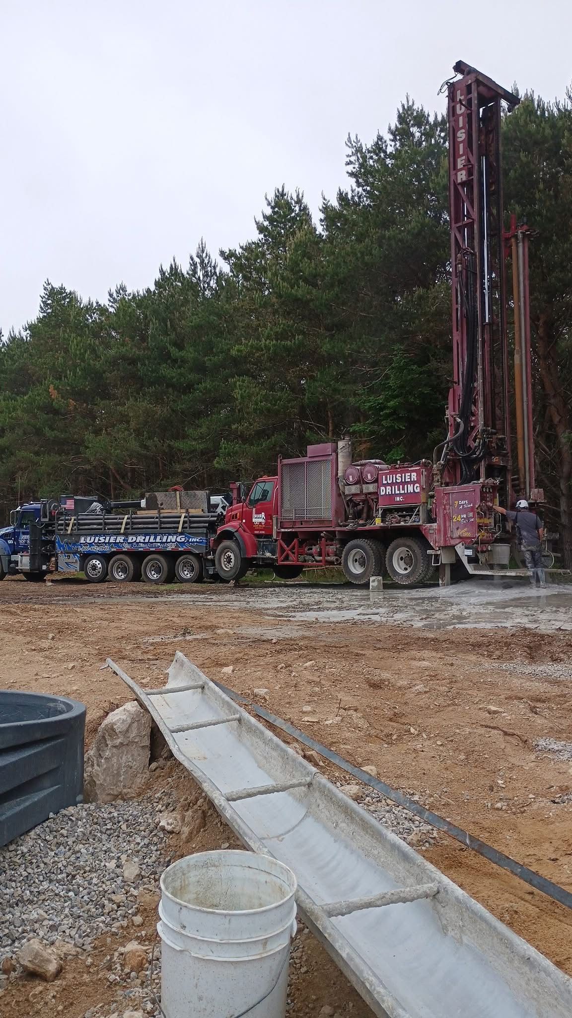 Drilling rig and truck on a muddy work site beside trees and a concrete drainage channel