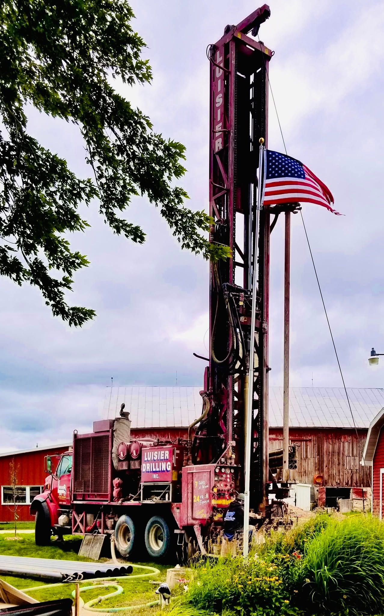 Red drilling rig truck beside a building, with a large American flag flying overhead.