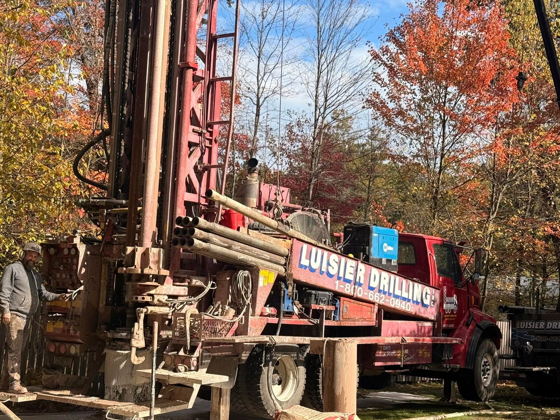 Drilling rig truck in a wooded autumn setting, with a worker nearby.